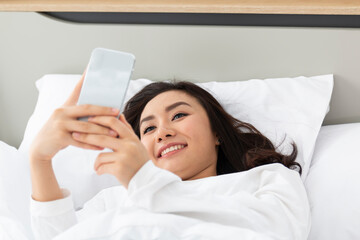 Obraz na płótnie Canvas Female playing with smartphone while laying on bed. Asian girl relaxing on bed and playing with the mobile phone. Young Asian girl looking at her smartphone while laying on the bed.