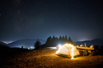Fantastic view of night starry sky over mountains with illuminated touristic tent. Back view of...