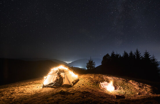 Fantastic view of starry sky over mountain valley with tourist tent and male traveler guitarist. Young man hiker sitting inside illuminated camp tent and playing guitar under night sky with stars.