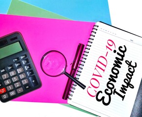 Covid-19 Economic Impact wording. Written on a note book with a magnifying glass, calculator and a pen on colorful A4 paper background.