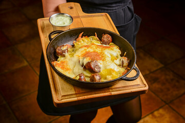 Sausages with herbs and cheese in a cast iron pan on a wooden board. Served by waiter in restaurant or diner.