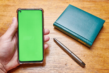 Male hand holds a phone with a green screen on a desktop background with a business notebook 2021 and a pen