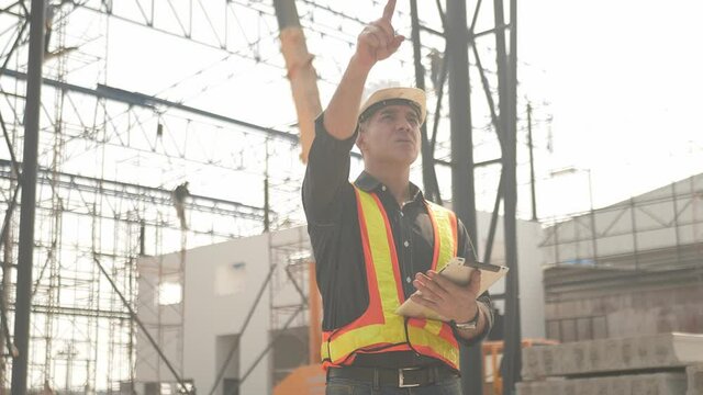 Foreman builder engineer worker using computer tablet looking at large building construction site sunny hot day hard work.