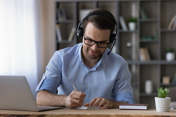 Smiling young Caucasian man in headphones sit at desk in home office study online on laptop making notes. Happy male in earphones have webcam digital virtual conference with client or customer.