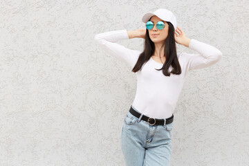 Portrait of beautiful adult woman with perfect body raising hands, wearing trendy jeans, shirt, baseball cap and sunglasses, looking away confident female posing against while wall.