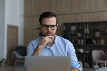 Young Caucasian man in glasses work online on computer in home office consult client customer distant. Pensive millennial male use laptop browse wireless Internet on gadget. Modern technology concept.