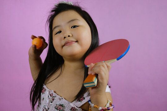 A cute young chubby Asian girl is showing off her ping pong paddle and ball, getting ready to play, feeling happy and confidence. Pink background.