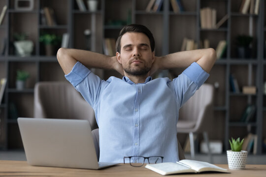Calm young Caucasian businessman sit lean in chair at workplace in home office breathe fresh condition air. Happy man relax rest at desk take nap or sleep, relieve negative emotions. Peace concept.