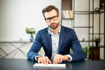 Serious elegant business man sitting in the modern spacious office typing on the white keyboard, looking directly at camera, chatting with business partners, managing large company remotely, big boss