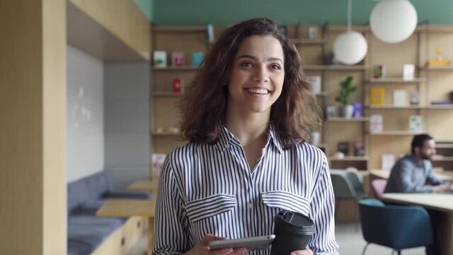 Happy latin girl student holding coffee and digital tablet walking in campus hallway greeting friends. Smiling hispanic female employee going along modern office coworking space.