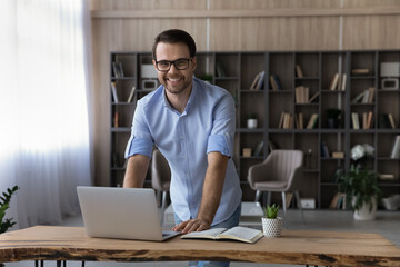 Portrait of smiling young Caucasian businessman stand at desk in home office work online on laptop gadget. Happy man use computer consult client or customer distant on device. Technology concept.
