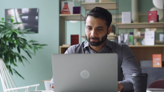 Young bearded indian businessman working on laptop in modern office lobby space. Indian executive using computer remote studying, watching online webinar, virtual training from homeoffice.