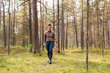 Fototapeta premium picking season and leisure people concept - happy smiling middle aged man with wicker basket of mushrooms walking in autumn forest