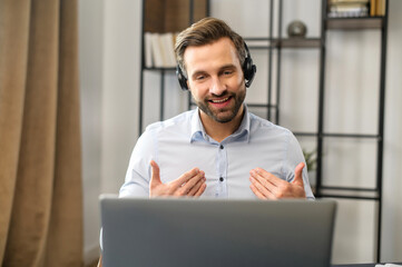 Positive concentrated millennial slavic-looking guy wearing earphones, listening to client while planning a relief strategy, focused young businessman takes part in an educational online conference