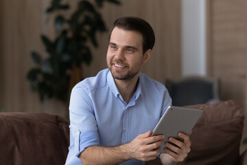 Happy millennial man relax at home use modern tablet gadget look in distance thinking planning. Smiling young Caucasian male hold pad device make decision. Technology, communication concept.