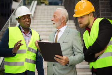 New plan. The construction site boss has a conversation with the architects. Focus is on background.