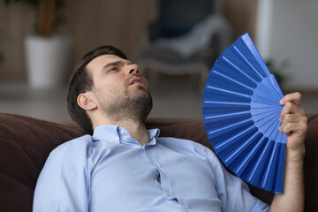 Unhappy tired young Caucasian man relax on sofa at home wave with hand fan suffer from heatstroke. Exhausted millennial male use waver breathe fresh air, struggle lack conditioner indoors. - obrazy, fototapety, plakaty