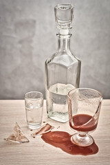 A broken glass, red tomato juice spilled on the table next to a bottle of vodka