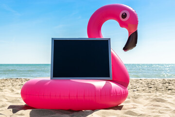 Flamingo icon. Funny pink toy flamingo with blackboard for text on summer ocean nature beach background in sunny tropical day. Pool float party. © Maksym