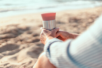 Woman's hand holding a thermos cup by autumn sea. Morning picnic with thermos of hot tea or coffee...