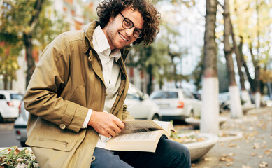 Horizontal image of a young man reading a book outdoors. College male student learns on campus in...