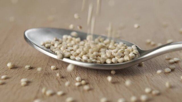 Raw quinoa seeds fall into a metal spoon in slow motion on a wooden board. Macro shot