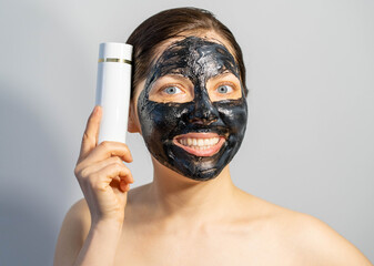 Emotional Woman with black charcoal face mask and white tube of cream in her hands