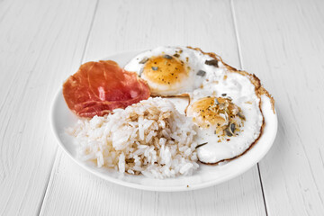 Two fried eggs sprinkled with dry green onions and a piece of bacon and rice topped with soy sauce for a side dish. Proteins, carbohydrates, fats are a balanced meal for lunch.