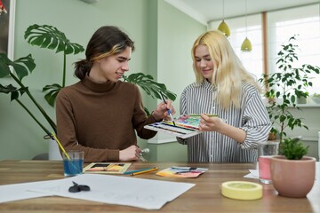 Teenagers guy and girl sitting at table with paints, brushes, drawings