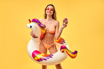 Cute woman in swimsuit with unicorn inflatable multicolored circle and pink donut isolated on yellow background. Summer time. Rest, vacations, travel.