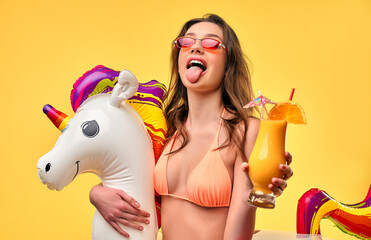 Cute woman in swimsuit with unicorn inflatable multicolored circle and cocktail isolated on yellow background. Summer time. Rest, vacations, travel.