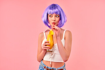 Beautiful young sexy girl in a purple wig drinks an ornamental cocktail with an orange isolated on a pink background. Summer time, rest, entertainment.