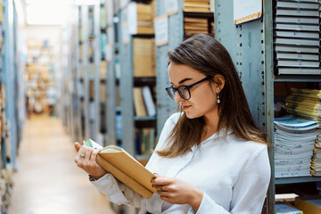 Diligent female student in a white blouse and glasses reading a book in the library. Concept of...
