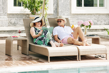 Mature couple reading and drinking cocktails when relaxing on chaise-lounges by swimming pool