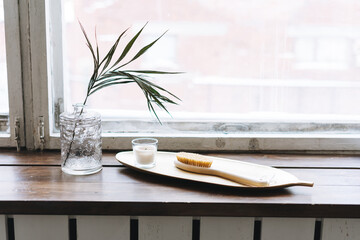 Details of interior, aromatic candle in glass and wooden brush with natural bristles on metal tray on the wooden window sill