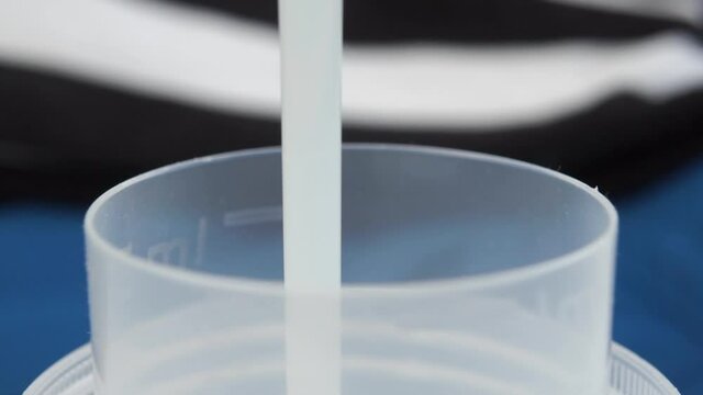 A stream of white liquid laundry detergent is poured into a plastic measuring cap against the background of a pile of laundry. Slow motion. Macro shot. Household chores concept