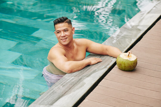 Cheerful middle-aged man standing in swimming pool of spa hotel and drinking sweet delicious coconut cocktail