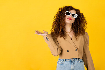 Curly girl in the trendy outfit of jeans and jacket posing on the yellow background in the studio. Woman facial expression. Isolated portrait.