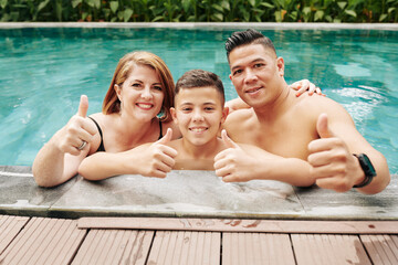 Cheerful excited father, mother and son standing in swimming pool and showing thumbs-up