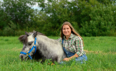 Happy young woman is sitting on the grass and hugging her horse.