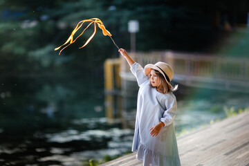 Child girl 5, 6, 7 years old in wicker hat and dress on shore of lake, evening light in summer and...