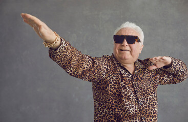 Retired senior man vibing to hype pop music. Portrait of funny rich white haired old grandpa in cool glasses and leopard patterned party shirt flexing, dancing and having fun on gray studio background