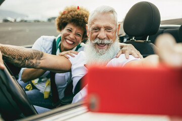 Trendy senior couple having fun in convertible car during in summer vacation - Joyful elderly people taking selfie on cabriolet auto outdoor with mobile phone - Powered by Adobe