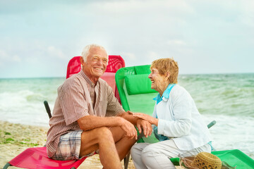 A very old married couple laughs cheerfully, sitting on sun loungers by the sea.