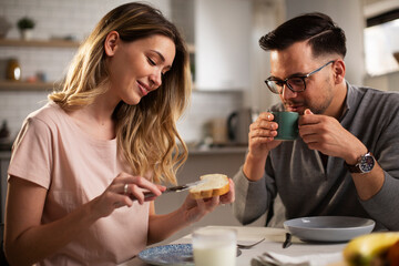 Beautiful young woman enjoying in breakfast with boyfriend. Happy young couple drinking coffee and eating sandwich at home..