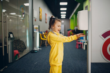 Child girl using automatic alcohol gel dispenser spraying on hands sanitizer machine antiseptic...