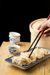 Top view of woman's hand with chopsticks holding a gyoza on blue plate, on wooden table with soy bowl and steamer, black background, with copy space