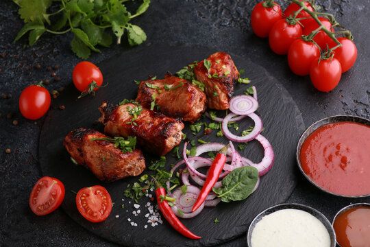 Meat and barbecue. Fried pieces of pork with fresh herbs, cilantro, onions, peppers, spices and vegetables on a black table. Assorted sauces in a bowl. Background image, copy space