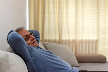 Obraz premium Peaceful older man feel relax on comfortable sofa in living room, calm mature male rest on cozy sofa, enjoy weekend at home. Side view of a 70s man with hands behind head enjoy at home, leaned on sofa