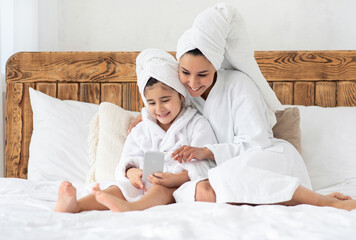 Beautiful mother and daughter in bathrobes using mobile phone